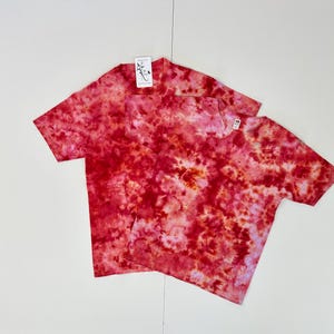 May include: Two short-sleeved t-shirts with a tie-dye pattern in shades of red, pink, and orange. One shirt has a pocket on the chest. Both shirts have tags attached. The shirts are on a white background.