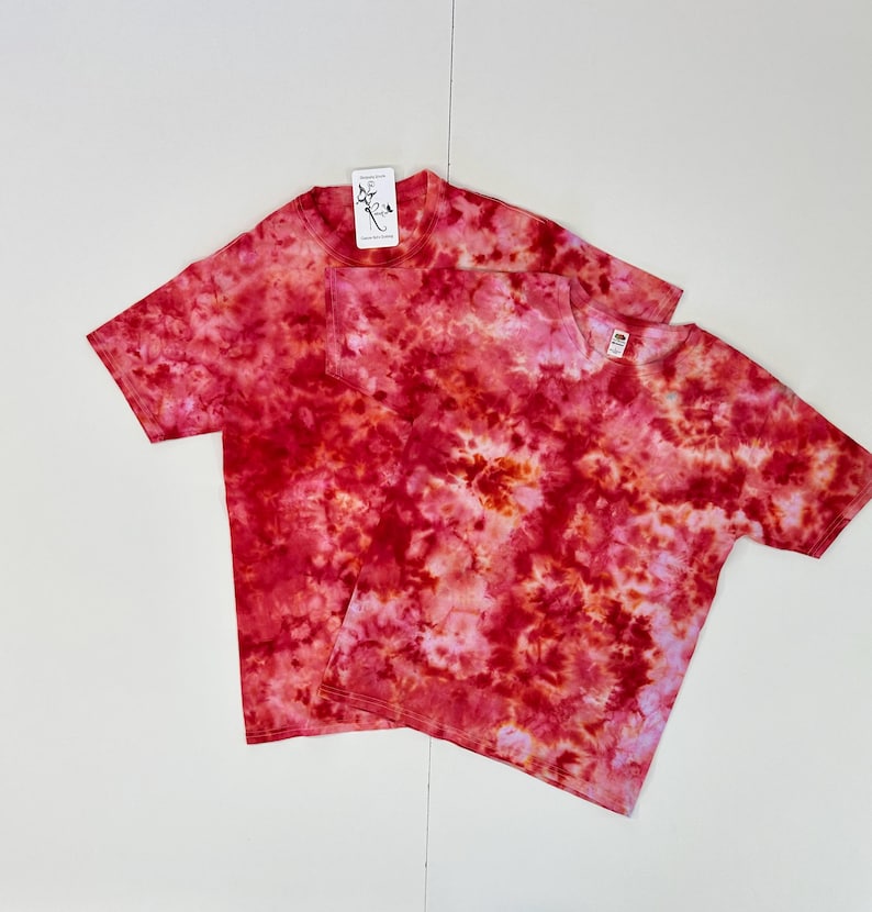 May include: Two short-sleeved t-shirts with a tie-dye pattern in shades of red, pink, and orange. One shirt has a pocket on the chest. Both shirts have tags attached. The shirts are on a white background.