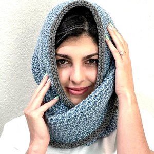 以下が含まれることがあります： 青いグレーのインフィニティスカーフは、質感のあるニットパターンでできています。スカーフは人が着用しており、折り返してタートルネックになっています。