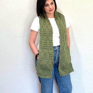 K&ouml;nnte beinhalten: Ein gestrickter gr&uuml;ner Schal mit Taschen. Der Schal hat eine olivgr&uuml;ne Farbe und wird &uuml;ber einem wei&szlig;en T-Shirt und einer blauen Jeans getragen. Der Schal ist aus einem dicken, strukturierten Garn gefertigt.