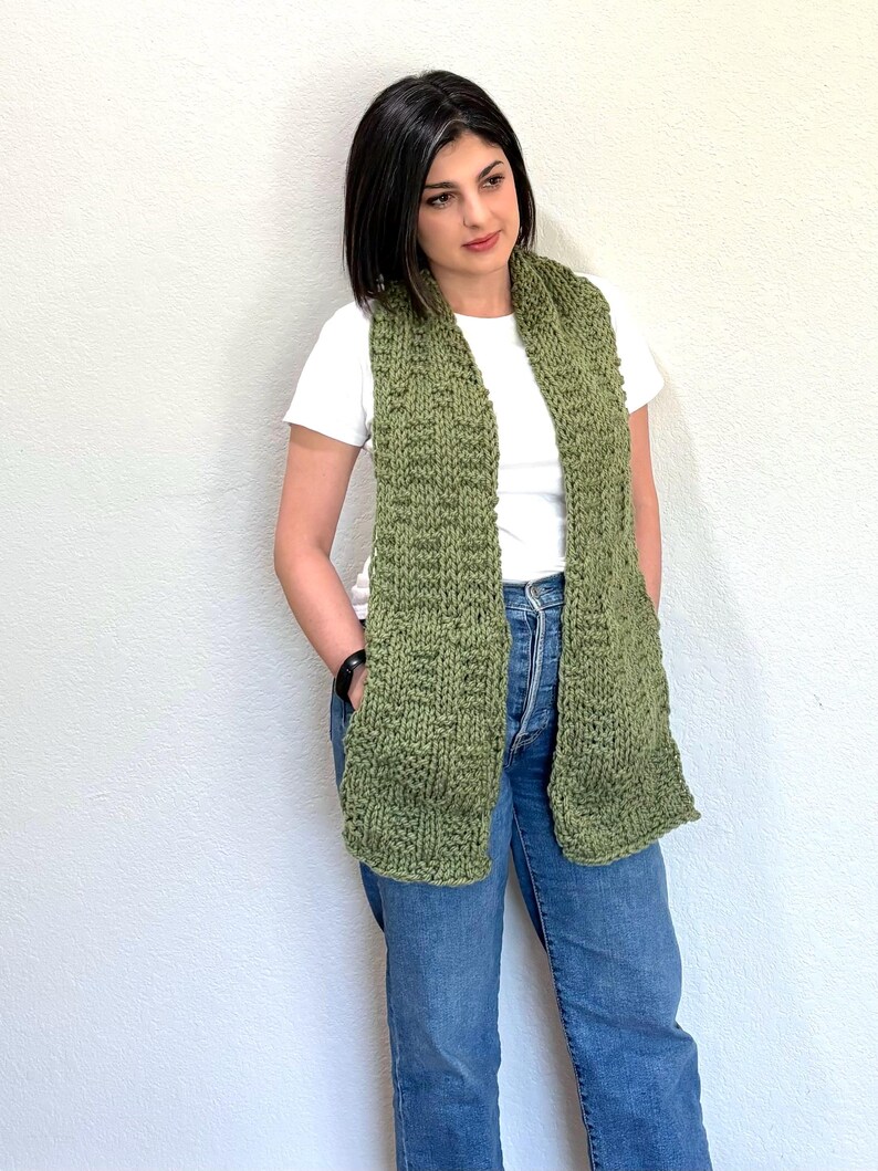 K&ouml;nnte beinhalten: Ein gestrickter gr&uuml;ner Schal mit Taschen. Der Schal hat eine olivgr&uuml;ne Farbe und wird &uuml;ber einem wei&szlig;en T-Shirt und einer blauen Jeans getragen. Der Schal ist aus einem dicken, strukturierten Garn gefertigt.