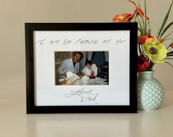 Puede incluir: Una foto enmarcada en negro con un mensaje escrito a mano que dice "Estoy tan orgulloso de ti" y "Love Dad". La foto muestra a un hombre y una niña pequeña sentados en una mesa.