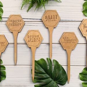May include: Six wooden plant markers with humorous sayings. The sayings are "Cheaper than Therapy", "Still Alive", "Proud Plant Mom", "She Bought Another One", "Thirsty AF", and "You Grow Girl".