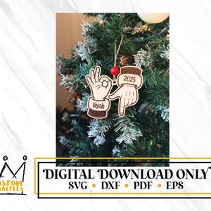 May include: A wooden Christmas ornament with a hand gesture design. The ornament features a hand making the "OK" sign and another hand making a gesture with the text "2025". The name "Nyah" is also on the ornament. The ornament is hanging on a Christmas tree.