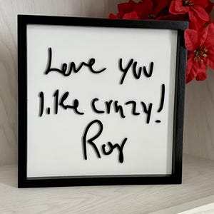 May include: A square sign with a black frame and white background, featuring black handwritten text that reads "Love you like crazy! Roy"