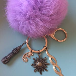 May include: A purple fluffy keychain with a gold ring and several charms, including a tower, a sun, a lizard, and a swirl design.