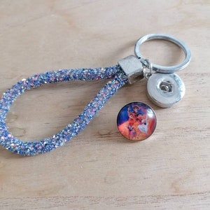 Könnte beinhalten: Ein blauer Glitzer-Schlüsselanhänger mit einem silbernen Ring und zwei Druckknöpfen. Ein Knopf zeigt ein farbenfrohes Bild von Figuren aus dem Film Coco.