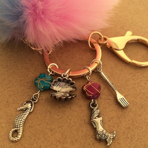 May include: A pink and blue fuzzy keychain with a gold ring and charms. The charms include a seashell, a mermaid, a seahorse, a red and gold bead, and a blue and gold bead.