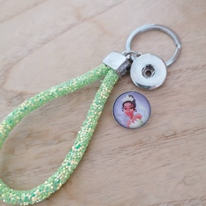 May include: A green glitter keychain with a silver ring and a snap button charm featuring a cartoon image of Princess Tiana from Disney's The Princess and the Frog.