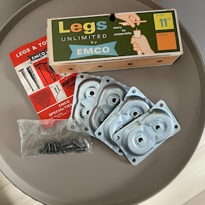 Puede incluir: Un conjunto de placas de montaje de patas de muebles de metal plateado, tornillos y una caja de 28 cm de "Legs Unlimited" de EMCO. La caja es verde, amarilla y blanca, con una ilustración de las instrucciones de montaje. También se incluye un folleto rojo y blanco.