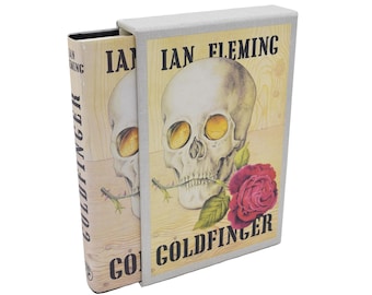 May include: A hardcover book titled "Goldfinger" by Ian Fleming, featuring a skull with golden eye sockets and a red rose. The book is in a light-colored slipcase.
