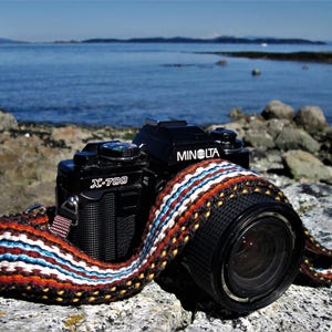 Könnte beinhalten: Eine schwarze Minolta X-700 Filmkamera mit einem bunten gewebten Kameragurt, die auf einer felsigen Oberfläche in der Nähe eines Gewässers liegt.