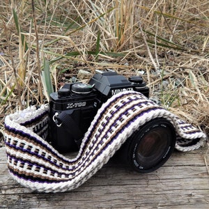 Peut inclure: Un appareil photo noir Minolta X-700 avec une sangle tissée. La sangle présente un motif répété de rayures blanches, marron et violettes. L'appareil photo repose sur une surface en bois vieilli, avec de l'herbe sèche en arrière-plan.