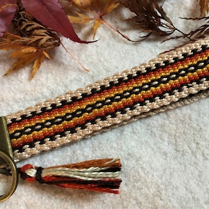 May include: A woven keychain with a black, orange and yellow pattern and a brown tassel. The keychain has a metal ring and a metal clasp.