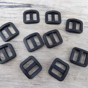 May include: Ten black plastic slider buckles. The buckles are square shaped with two parallel slots in the centre.