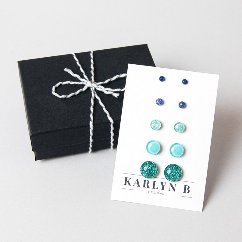 May include: A set of six pairs of stud earrings in a black gift box. The earrings are in shades of blue and green, with some having glitter. The card reads "KARLYN B DESIGNS".