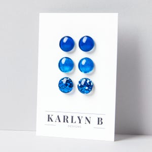 May include: Three pairs of circular stud earrings displayed on a white card labeled KARLYN B DESIGNS. The top pair is solid cobalt blue, the middle pair features a shimmering blue finish, and the bottom pair is filled with chunky blue glitter.