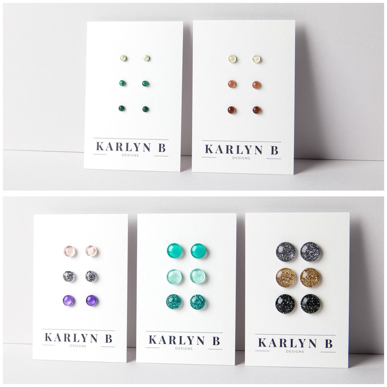 May include: Five cards with the text 'KARLYN B DESIGNS' printed on them. Each card features a set of three stud earrings with different colors and designs. The earrings are arranged in a vertical line on each card. The colors of the earrings include green, white, brown, pink, purple, teal, gold, and black.