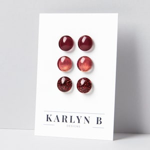 Op de afbeelding: Drie paar ronde oorknopjes gepresenteerd op een witte kaart met de tekst KARLYN B DESIGNS. Het bovenste paar is effen donkerrood, het middelste paar is glinsterend koperrood en het onderste paar heeft een dieprode basis met fijne glitters.