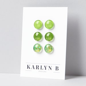 May include: Three pairs of round stud earrings displayed on a white card labeled KARLYN B DESIGNS. The top pair is solid light green, the middle pair is a darker translucent green, and the bottom pair features a multi-colored glitter finish.