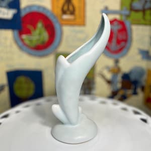 May include: A light blue ceramic bud vase with a curved, organic shape. The vase has a flared opening and a small, rounded base. The interior of the vase is a lighter shade of blue. The vase is sitting on a white, decorative surface.