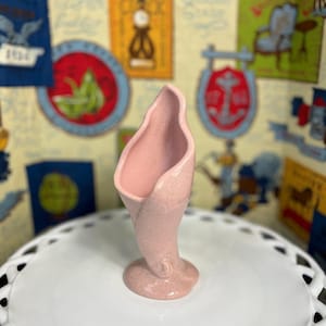 May include: A light pink ceramic vase with a speckled texture. The vase has a unique, curved shape, resembling a calla lily. It sits on a round base and is displayed on a white, decorative plate.