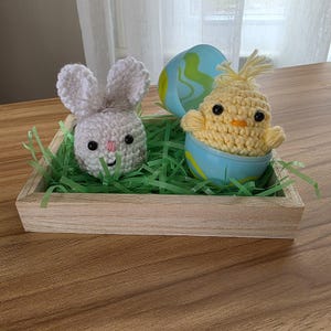 May include: A wooden tray filled with green paper grass, featuring a crocheted Easter bunny and a chick in an egg. The bunny is white with pink accents, and the chick is yellow. An eggshell is behind the chick.