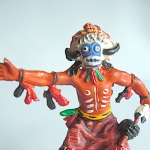 Può includere: Una statuetta di plastica di un ballerino nativo americano con una maschera di teschio di bufalo, abiti rossi e gialli e un copricapo piumato. La statuetta è in piedi su una base verde.
