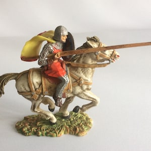May include: A miniature figurine of a knight on horseback, charging with a lance. The knight is wearing chainmail armor, a red tunic, and a yellow cape. The horse is white with brown tack.