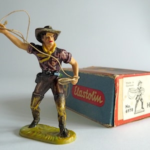 May include: A plastic toy cowboy figure with a brown hat, purple shirt, and brown trousers, holding a lasso. The figure is standing on a green base. The figure is packaged in a blue and red box with the text "Elastolin" and "No. 6978" printed on it.