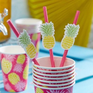 May include: Three pink paper straws with pineapple designs on the top. The straws are in a stack of pink and white paper cups with pineapple designs.