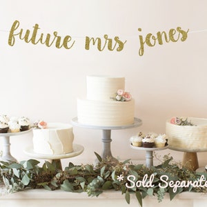 May include: Gold glitter banner with the text "future mrs jones" hanging above a table with two white cakes, cupcakes, and greenery. The text "Sold Separately" is written below the table.