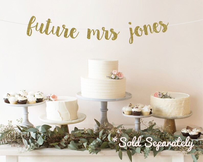 May include: Gold glitter banner with the text "future mrs jones" hanging above a table with two white cakes, cupcakes, and greenery. The text "Sold Separately" is written below the table.