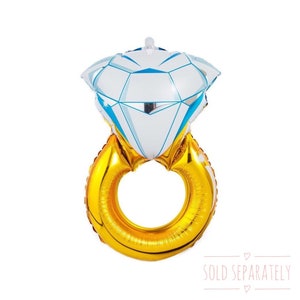 May include: A gold ring balloon with a large white diamond balloon on top. The balloons are shaped like a ring and diamond. The text "SOLD SEPARATELY" is printed below the balloons.