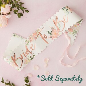 May include: White satin sash with a scalloped edge and a rose gold foil inscription that reads "Bride To Be". The sash is decorated with a floral pattern in shades of pink and green.