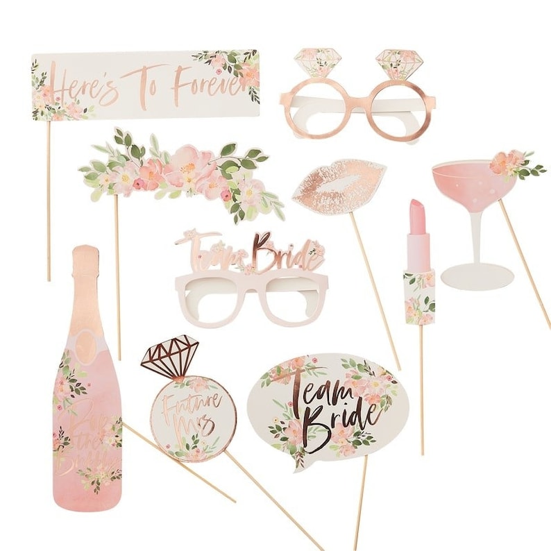 May include: A set of 10 pink and gold floral themed photo booth props for a bridal shower or bachelorette party. The props include a champagne bottle, a lipstick, a ring, a speech bubble that says "Team Bride", a pair of glasses that say "Forever Bride", a pair of glasses with rings, a pair of lips, a champagne glass, and a sign that says "Here's To Forever".