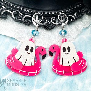 Op de afbeelding: Dangle oorbellen met een witte geest op een roze flamingo zwemband. De oorbellen hebben zilverkleurige haken en blauwe kraalaccenten. De oorbellen zijn geplaatst tegen een decoratieve zwarte achtergrond en een blauw waterachtig oppervlak. De tekst "Sparkle Monster" is zichtbaar.