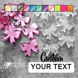 May include: Assortment of clear and pink acrylic bow shapes with the text "sparkle monster" printed on them. The bows are available in a variety of colours and can be customised with your own text.