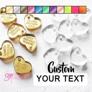 May include: Assortment of gold and clear acrylic heart-shaped charms with the text "sparkle monster .com" printed on them. The charms are scattered on a white surface. The text "Custom Your Text" is printed below the charms.
