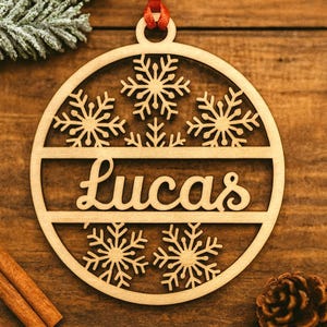 May include: Wooden Christmas ornament with the name "Lucas" cut out in a cursive font. The ornament features snowflake designs and a red ribbon for hanging. The background is a wooden surface.