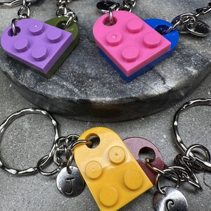 May include: Keyrings with heart-shaped Lego brick designs in colours such as purple, green, pink, blue, yellow, and maroon. Each keyring has a silver ring and a small, round charm with a letter.