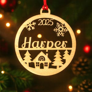 May include: A wooden Christmas ornament with a red ribbon. The ornament is circular with the name "Harper" and the year "2025" cut out. It features snowflake and tree designs, and a house silhouette.
