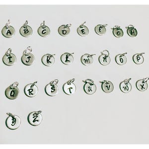 May include: A selection of small, silver-coloured alphabet charms, each with a loop for attaching to a necklace or bracelet. The letters A to Z are displayed in a clear, readable font. These charms are ideal for personalised jewellery.