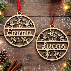 May include: Two wooden Christmas ornaments with snowflake designs and the names "Emma" and "Lucas" are displayed. Each ornament hangs from a red ribbon. The background includes pine needles, pine cones, and cinnamon sticks.