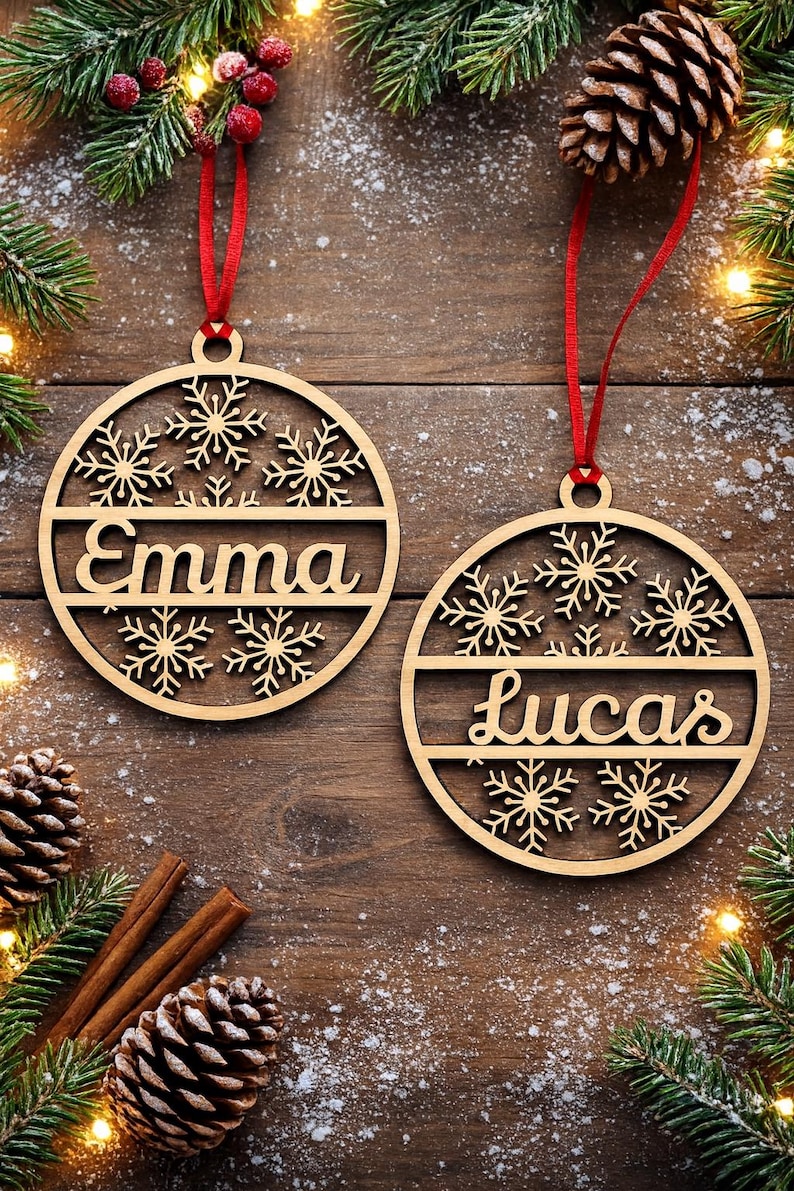 May include: Two wooden Christmas ornaments with snowflake designs and the names "Emma" and "Lucas" are displayed. Each ornament hangs from a red ribbon. The background includes pine needles, pine cones, and cinnamon sticks.