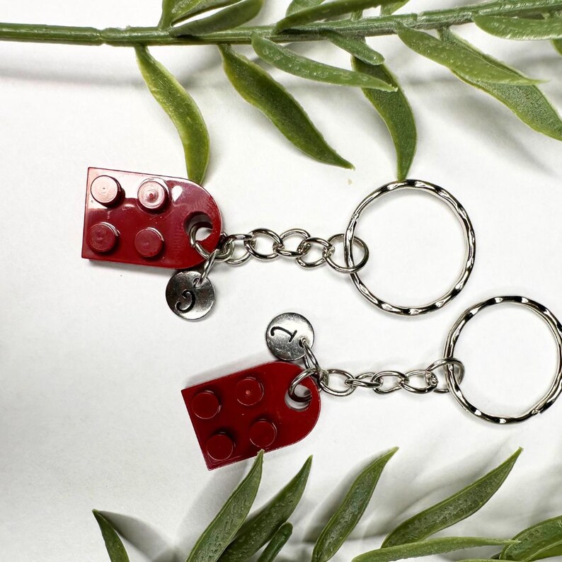 May include: Two red brick-shaped keyrings with silver key rings. Each keyring has a small silver charm with a letter. The keyrings are connected by a silver chain. Green leaves are in the background.