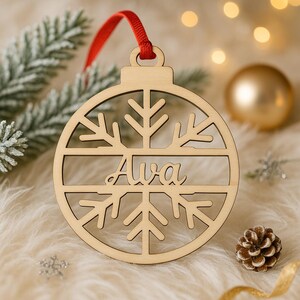 May include: Wooden Christmas ornament with a snowflake design and the name "Ava" cut out. The ornament hangs from a red ribbon. The background includes a pine branch, a gold ornament, and a pine cone.