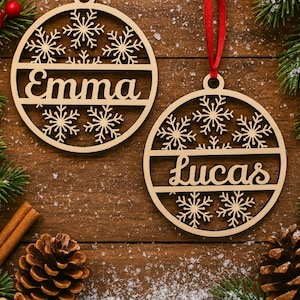 May include: Two wooden Christmas ornaments with snowflake designs and the names "Emma" and "Lucas" cut out. The ornaments hang from red ribbons. The background includes pine needles, cinnamon sticks, and pine cones on a wooden surface.