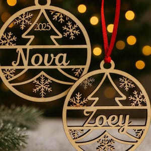 May include: Two wooden Christmas ornaments with red ribbons. One ornament says "Nova" and "2025" with a tree and snowflake design. The other ornament says "Zoey" with a tree and snowflake design.
