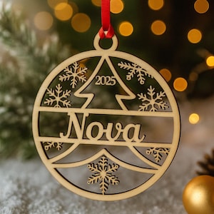May include: Wooden Christmas ornament with the name "Nova" and the year "2025" cut out. The ornament features a tree and snowflake design, suspended by a red ribbon. The background is blurred with Christmas lights.